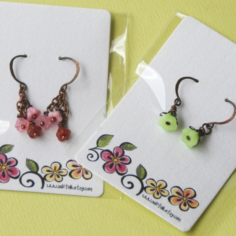 May include: Two pairs of earrings displayed on white cards with floral designs. One pair features pink flower-shaped beads and the other pair features green flower-shaped beads. Both pairs have copper-coloured hooks. The cards have the website www.selftakest.etsy.com printed on them.