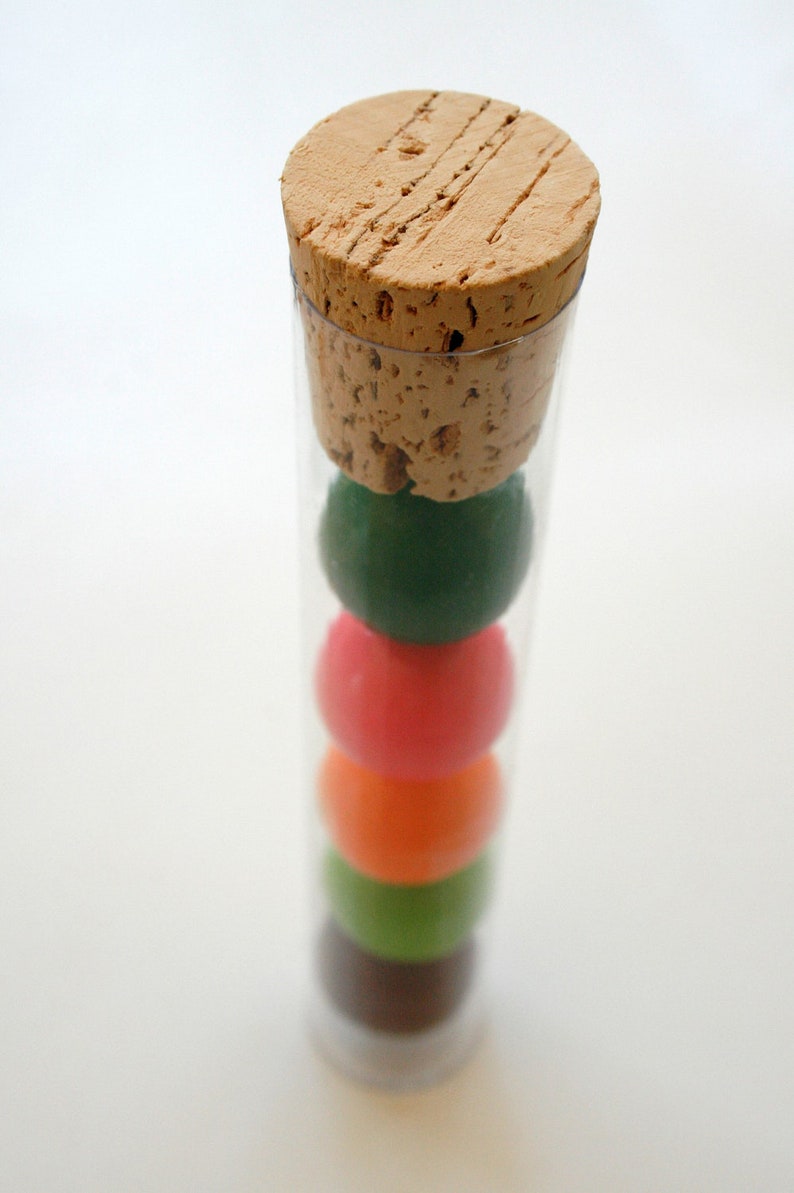 Pu&ograve; includere: Un tubo di vetro trasparente con un tappo di sughero e quattro palline colorate all'interno. Le palline sono verdi, rosa, arancioni e verdi.