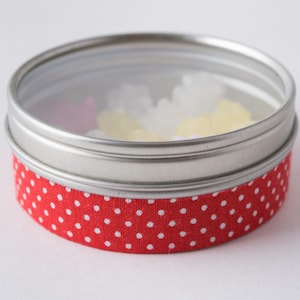May include: A round metal tin with a red and white polka dot fabric lid. The lid has a clear plastic window that shows colorful candies inside.