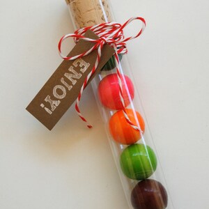 Pu&ograve; includere: Un tubo di vetro riempito con caramelle colorate, legato con uno spago rosso e bianco e un'etichetta marrone che dice "Enjoy!"
