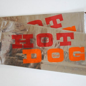 May include: Two silver foil hot dog wrappers with the word "HOT DOG" printed in red and orange letters. The top wrapper has the word "Frank's" printed in a smaller font and the bottom wrapper has the word "Delicious" printed in a smaller font.