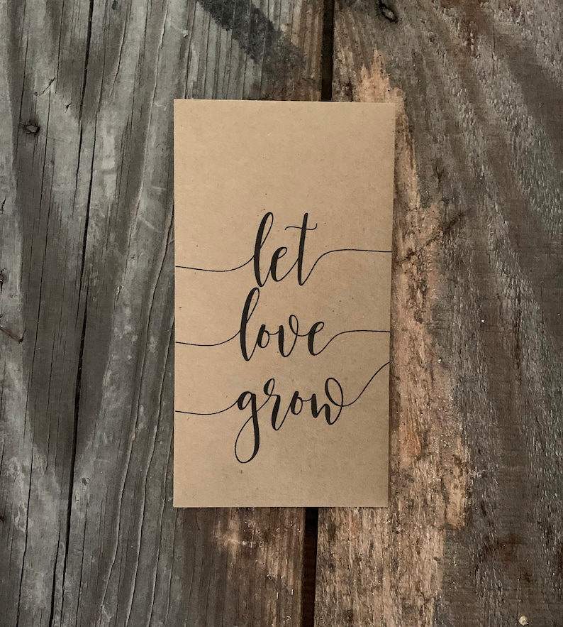 以下が含まれることがあります： 黒い筆記体で「let love grow」と書かれた茶色の紙のカードです。