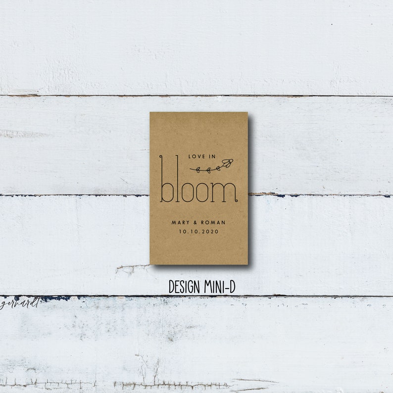 May include: A brown paper card with the text "Love in Bloom" and a simple floral design. Below the text is "Mary & Roman 10.10.2020". The card has the text "Design Mini-D" printed below it.