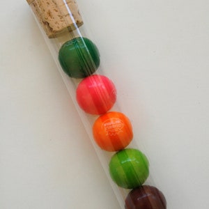 Pu&ograve; includere: Un tubo di vetro trasparente con un tappo di sughero riempito con cinque gomme da masticare colorate. Le gomme da masticare sono verdi, rosa, arancioni, verdi e marroni.