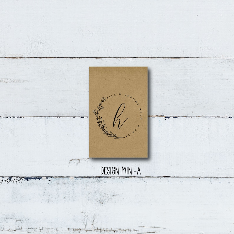 May include: A brown paper favor bag with a black and white design. The design features a floral wreath with the letter "H" in the center. The text "Jill & Jerome Helm 12.21" is written around the wreath.