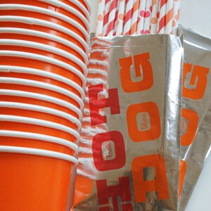 May include: A stack of orange and white paper cups with a silver foil bag with the word "HOT DOG" printed in red letters. The bag also has the words "Fresh" and "Delicious" printed in red letters.