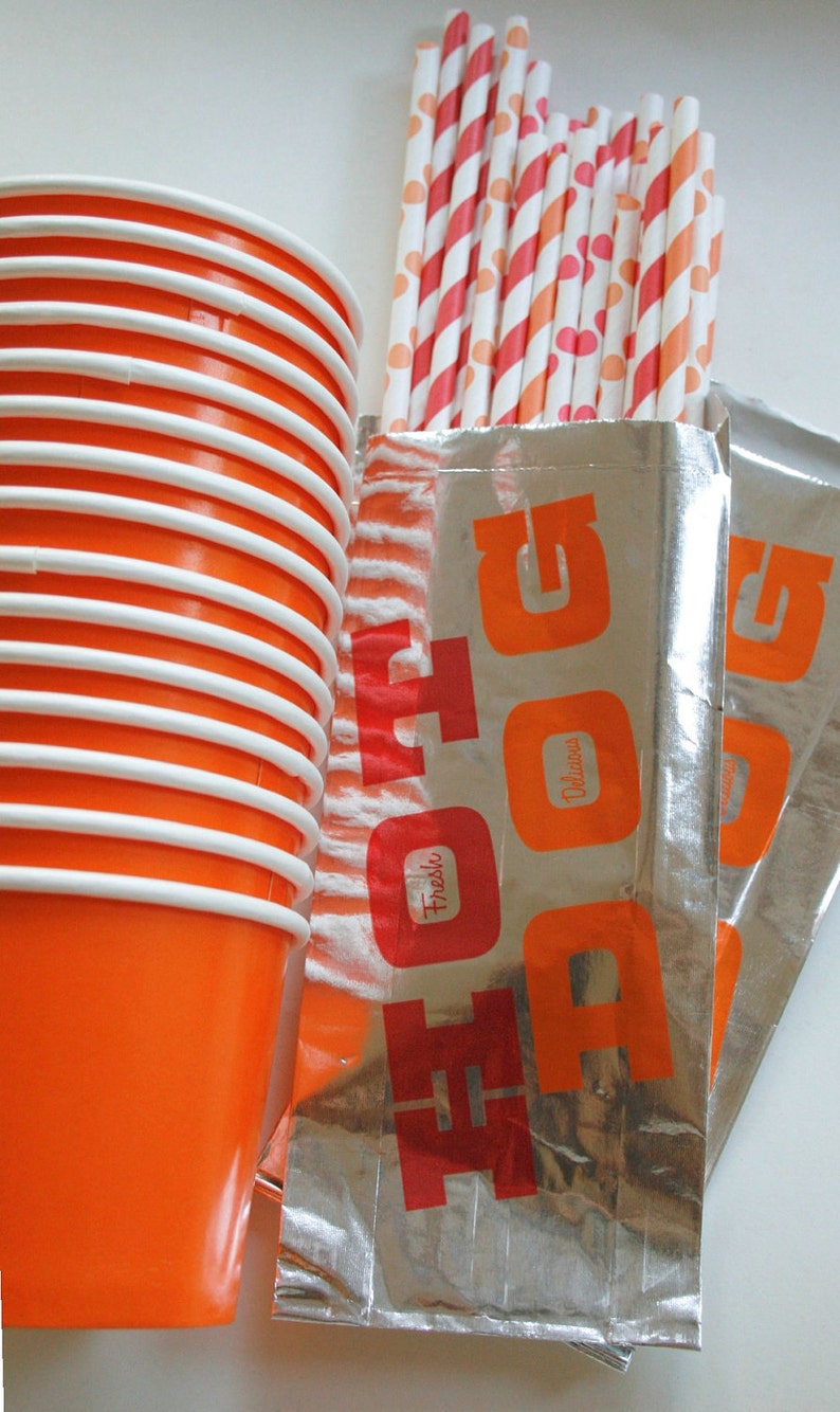 May include: A stack of orange and white paper cups with a silver foil bag with the word "HOT DOG" printed in red letters. The bag also has the words "Fresh" and "Delicious" printed in red letters.