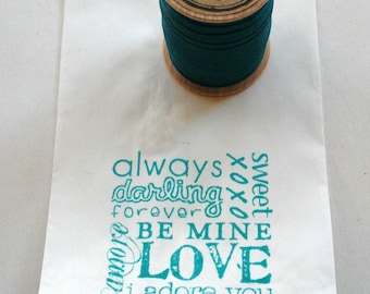 Puede incluir: Una bolsa de papel blanca con un sello azul turquesa que dice "always darling forever xoxo sweet BE MINE LOVE i adore you". Un carrete de hilo azul turquesa está en la parte superior de la bolsa.