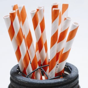 May include: A close-up of a bunch of orange and white striped paper straws in a black metal holder.