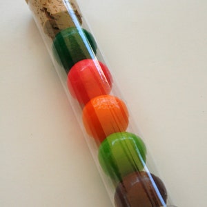 Pu&ograve; includere: Un tubo di vetro trasparente con un tappo di sughero riempito di cinque caramelle rotonde colorate. Le caramelle sono verdi, rosse, arancioni, verdi e marroni.