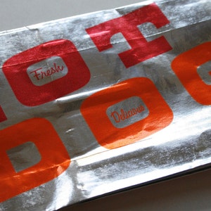 May include: Silver foil packaging with the words "Hot Dog" printed in red and orange letters. The words "Fresh" and "Delicious" are also printed in red and orange.