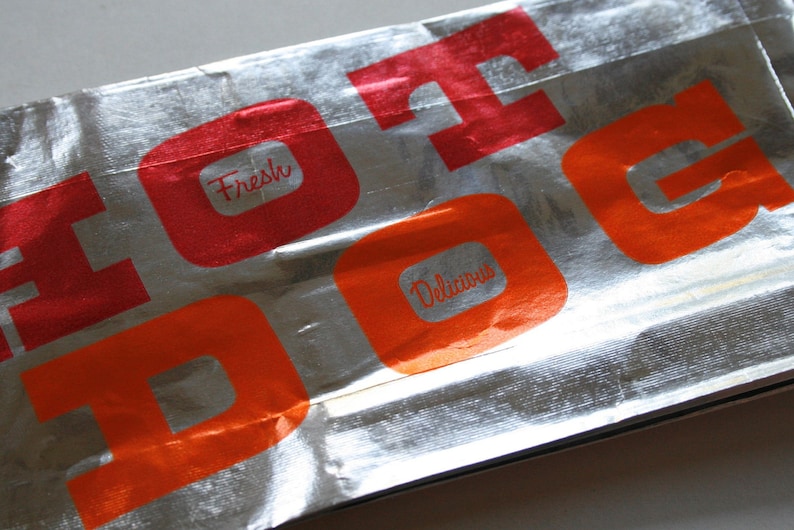 May include: Silver foil packaging with the words "Hot Dog" printed in red and orange letters. The words "Fresh" and "Delicious" are also printed in red and orange.