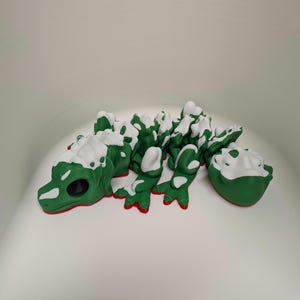 Puede incluir: Una figurita de lagarto verde y blanco con textura rugosa. El lagarto está acostado boca abajo con la cabeza girada hacia un lado. La figurita está hecha de un material suave y flexible.