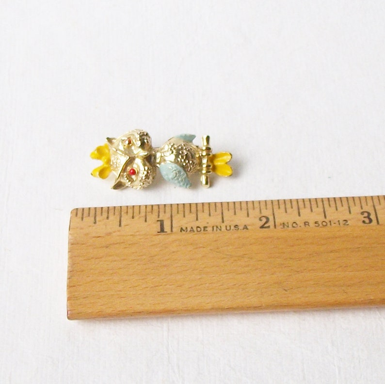 May include: A gold-tone owl brooch with red eyes, yellow accents, and a blue body. The brooch is approximately 2.5 cm long and is sitting on a ruler.