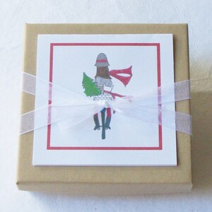 May include: A brown gift box with a white ribbon and bow. The box has a white card attached to the front with a red border. The card features a drawing of a person riding a bicycle with a Christmas tree in the basket.