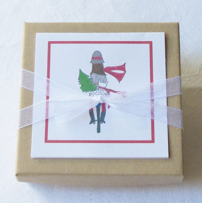 May include: A brown gift box with a white ribbon and bow. The box has a white card attached to the front with a red border. The card features a drawing of a person riding a bicycle with a Christmas tree in the basket.