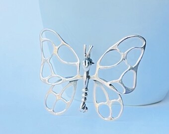 May include: A silver butterfly brooch with a delicate, openwork design. The wings are made of small, interconnected circles, creating a lacy effect. The butterfly's body is slender and detailed.