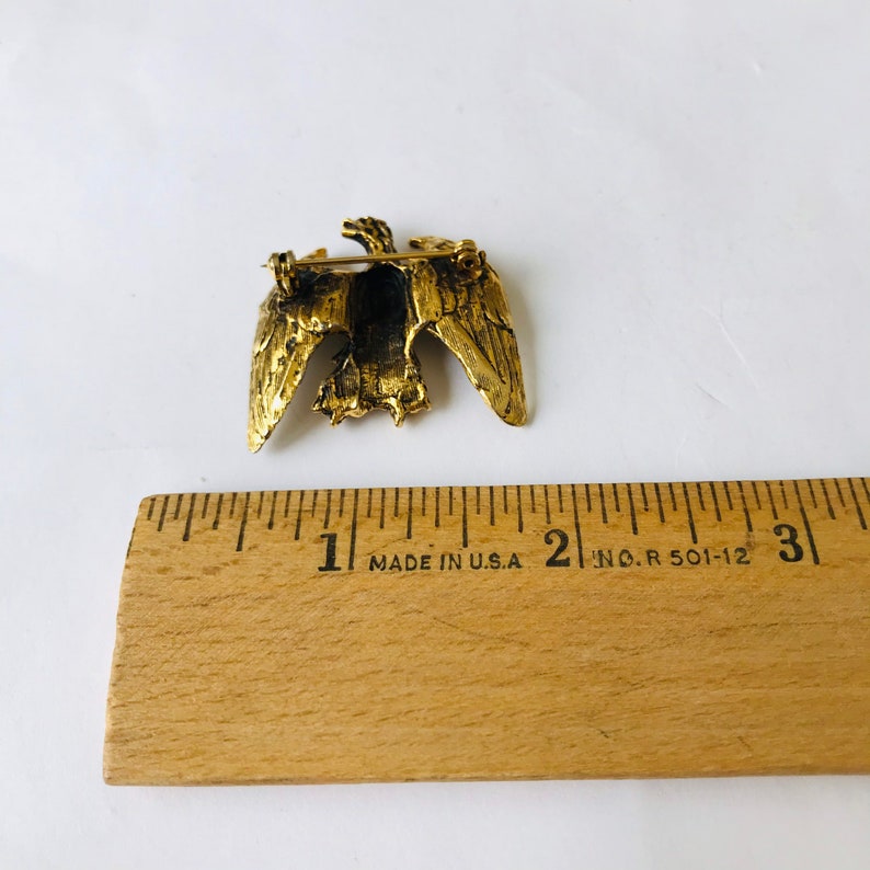 May include: A gold-toned metal eagle brooch with outstretched wings. The brooch is approximately 2 inches wide. The pin is made in the USA and is set against a white background.