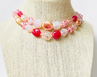 May include: A multi-strand necklace with pink, white, and clear oval beads. The necklace is made of glass and has a layered look.