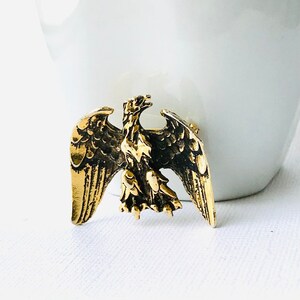 May include: A gold-colored metal eagle brooch with outstretched wings. The eagle is detailed with textured feathers and a detailed head. The brooch is likely vintage or antique, and is a decorative accessory.