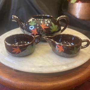 May include: A brown ceramic tea set with a teapot, creamer, and cup. The set is decorated with orange, green, and white leaf designs. The tea set is displayed on a white marble tray.
