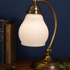 May include: A classic desk lamp with a brass finish. It has a curved arm and a white, ribbed glass shade. The base is circular, and the lamp sits on a wooden surface. Books and a pen are in the background. The lamp is reminiscent of a library.