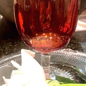 May include: A red glass goblet with a white rose and green leaves on a clear glass plate.