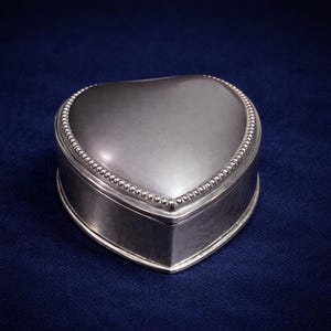 Puede incluir: Una caja de baratijas plateada en forma de corazón con un borde de cuentas. La caja tiene una superficie lisa y reflectante y una tapa con bisagras. La caja se muestra sobre un fondo azul oscuro.