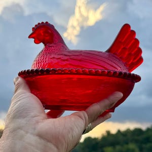 Può includere: Un piatto in vetro rosso vivace a forma di gallina su un nido. Il coperchio è modellato come una gallina seduta con texture di piume dettagliate, mentre la base presenta un bordo perlato e un motivo a cestino intrecciato in vetro rosso.