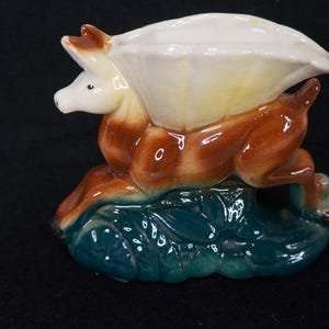 Puede incluir: Figura de cerámica de un ciervo saltando sobre un tronco estilizado. El ciervo es marrón y crema, con un objeto blanco en forma de concha en la espalda. La base es turquesa con detalles de hojas. Un artículo decorativo.