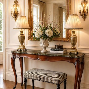 May include: An elegant entryway scene featuring a dark wood console table with a marble top, adorned with two lamps and a floral arrangement. A striped bench sits below. A large gold-framed mirror and sconces complete the classic decor.