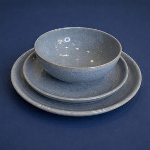 May include: A three-piece ceramic dinnerware set in a speckled blue-gray finish. The set includes a bowl and two plates of varying sizes, all with a slightly raised rim. The pieces are arranged on a dark blue surface, creating a cohesive and modern aesthetic.