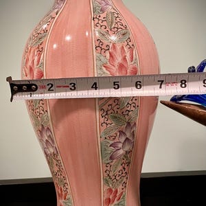 May include: A pink ceramic vase with floral designs and a brass base. The vase is approximately 7 inches wide. The floral designs include red, purple, and green lotus flowers. The vase has a glossy finish and a decorative shape.