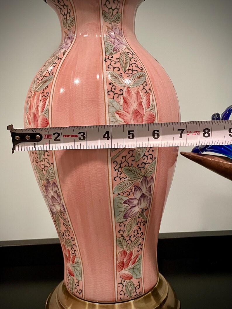 May include: A pink ceramic vase with floral designs and a brass base. The vase is approximately 7 inches wide. The floral designs include red, purple, and green lotus flowers. The vase has a glossy finish and a decorative shape.