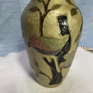 Puede incluir: Un pequeño jarrón decorativo de latón con un diseño floral dorado y verde y un pájaro multicolor en una rama negra.