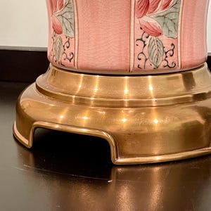 May include: A close-up of a pink ceramic lamp base with a gold-colored metal base. The lamp features a floral design with red and green accents. The lamp base has a textured surface.