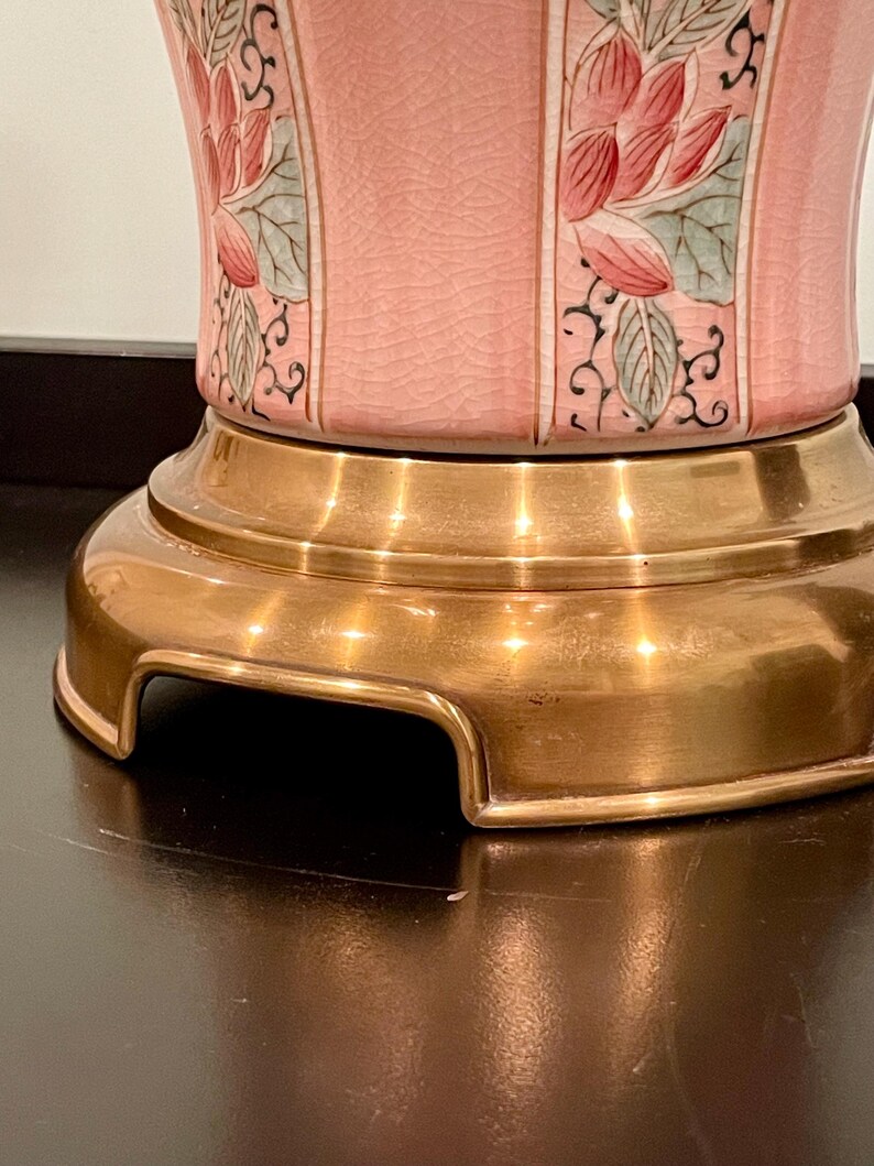 May include: A close-up of a pink ceramic lamp base with a gold-colored metal base. The lamp features a floral design with red and green accents. The lamp base has a textured surface.
