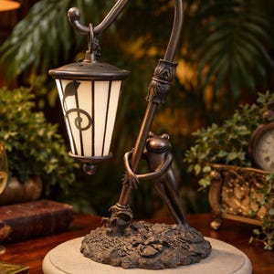 May include: A bronze-colored table lamp featuring a frog embracing the lamp post. The lamp has a white, rectangular shade with a decorative design. The lamp sits on a round, stone-like base. The lamp is a decorative home decor item.