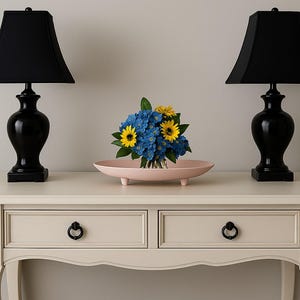 May include: A light pink oval-shaped decorative bowl with small feet holds a bouquet of blue and yellow flowers. Two black lamps with black shades flank the bowl on a cream-colored table.