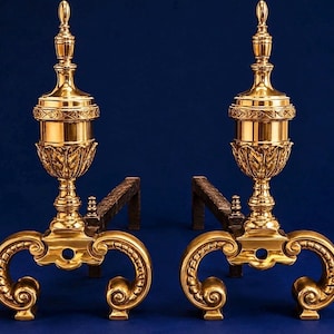 May include: A pair of polished brass fireplace andirons featuring ornate urn-shaped finials with leaf detailing, supported by curved scroll-work bases and extending into dark, textured iron horizontal bars for log support.