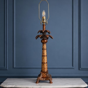 May include: A table lamp base shaped like a palm tree with a textured, light brown trunk and dark brown palm fronds at the top. The lamp features a brass-colored metal harp and socket assembly, standing on a square, decorative base with feet.