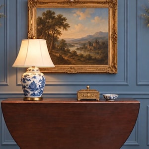 May include: A dark wood drop-leaf table featuring a blue and white porcelain lamp with a cream shade, a rectangular gold metal box with ornate detailing, and a small blue and white porcelain bowl, all set against a blue paneled wall.