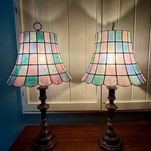 May include: Two antique table lamps with pastel-colored, stained-glass shades. The shades are divided into squares of pink, blue, and green. The lamps have ornate, dark bronze bases and are placed on a wooden surface.