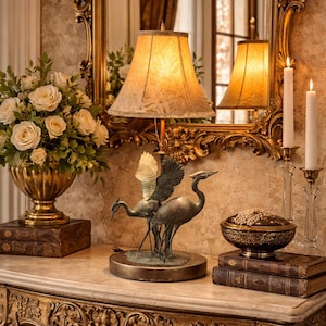 Könnte beinhalten: Ein goldfarbener Tisch mit kunstvollen Schnitzereien trägt eine Lampe mit gemustertem Schirm, eine Bronzekranichskulptur und ein Blumengesteck. Ein goldgerahmter Spiegel reflektiert die Lampe und die Kerzen.