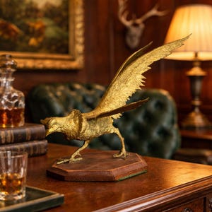 May include: A gold-colored bird sculpture with outstretched wings, mounted on a brown wooden base. The bird is detailed with textured feathers and stands on a hexagonal base. The sculpture is displayed on a wooden surface, with a glass of amber liquid and a decanter in the background.