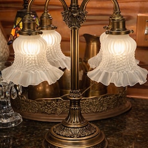 May include: An antique-style table lamp with three lights. The lamp features a bronze-colored base and stem with ornate detailing. Each light has a ruffled, frosted glass shade. The lamp is designed to provide ambient lighting.