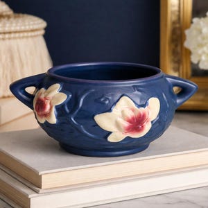 May include: A navy blue ceramic bowl with two handles and floral relief details. The bowl features cream-colored flowers with red centers. It sits atop two books with cream-colored pages.