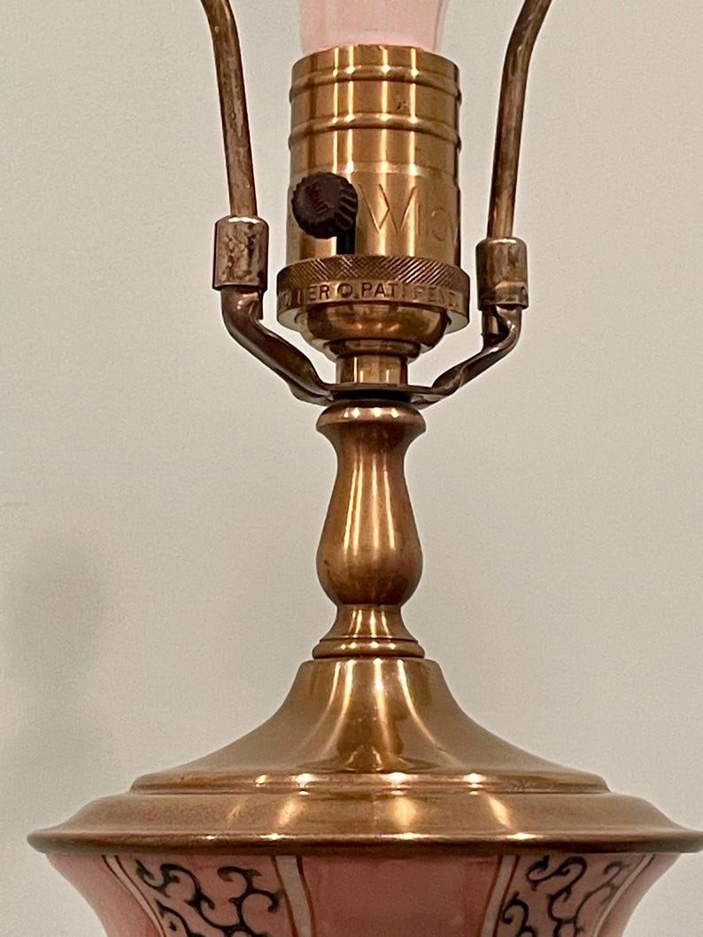 May include: Close-up of a vintage table lamp with a pink and black patterned base and a brass-colored metal body. The lamp has a bulb socket and a switch. The lamp is designed for indoor use.