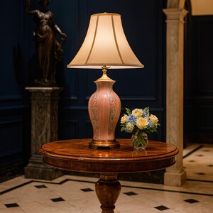 May include: A decorative table lamp with a beige lampshade and a pink base adorned with floral patterns. The lamp sits on a round, wooden table, accompanied by a small vase of flowers. The room has a dark blue wall and a statue in the background.