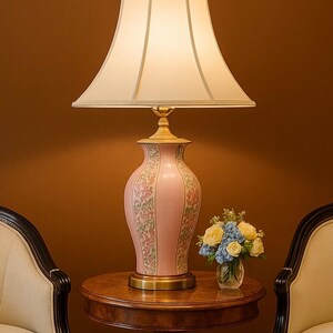 May include: A pink ceramic table lamp with a floral design, a cream-colored lampshade, and a brass base. The lamp sits on a round wooden table, next to a small vase of flowers. Two chairs are on either side of the table.